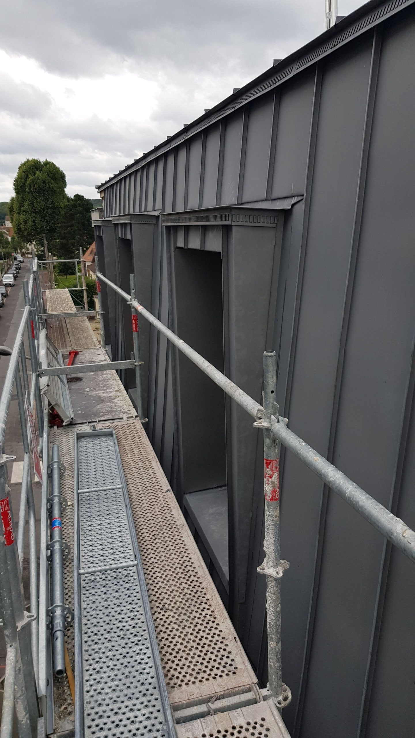 Lešenie popri streche modernej sivej kovovej budovy pod zamračenou oblohou. Kovové tyče a perforované lávky naznačujú prebiehajúci stavebný projekt.