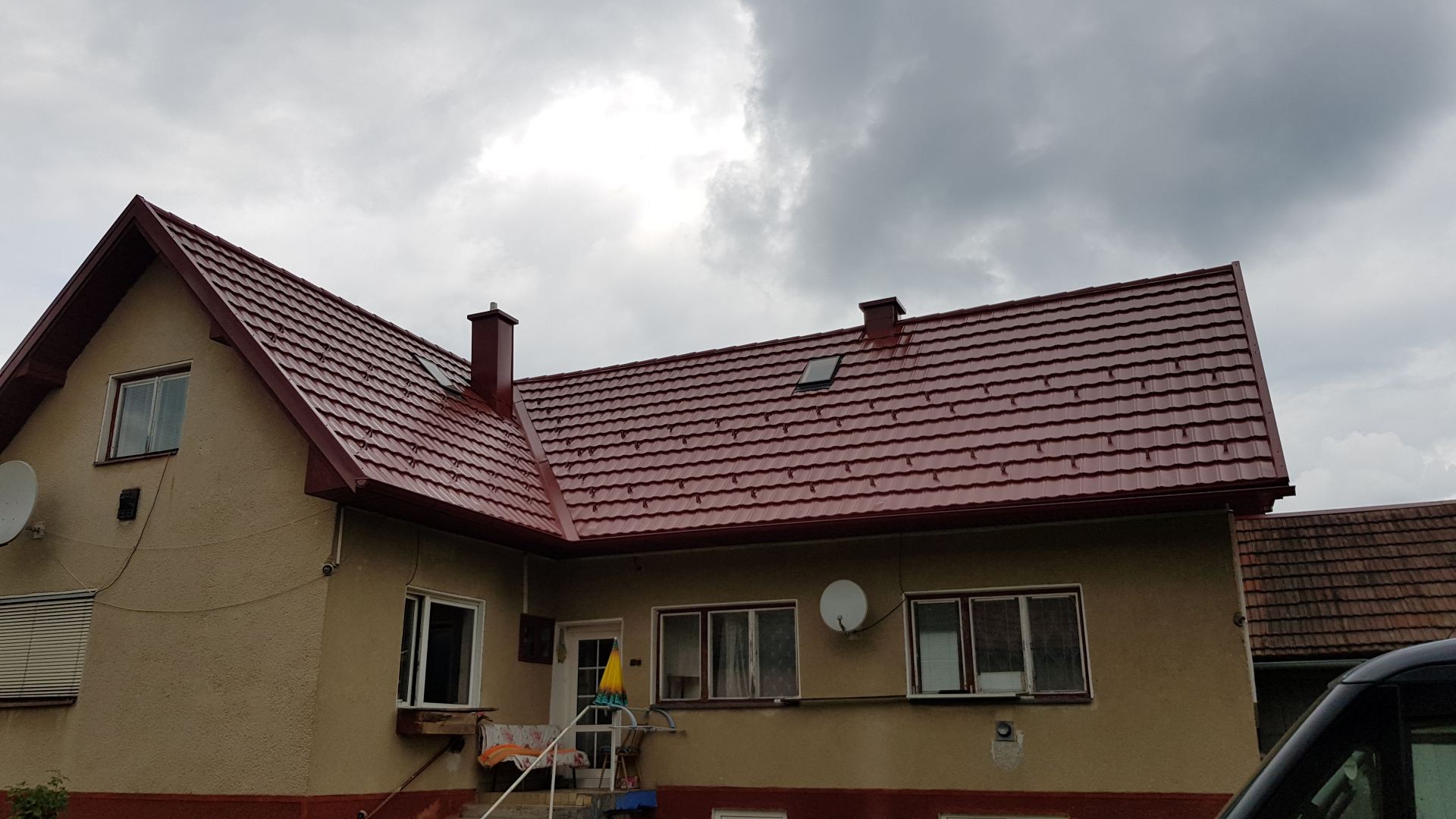 Dom so sedlovou strechou a červenými škridlami na pozadí zamračenej oblohy. Vidno béžové steny, okná s bielymi rámami a satelitnú anténu. Zamlžená a pokojná atmosféra.