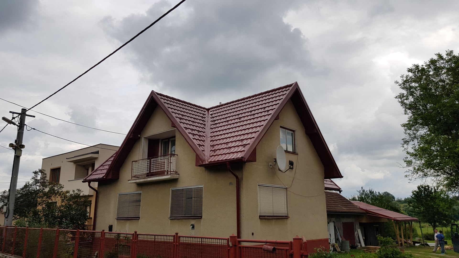 Béžový dom s červenou škridlovou strechou a satelitnou anténou pod zamračenou oblohou. Oplotená záhrada je lemovaná kríkmi a napravo stojí strom.