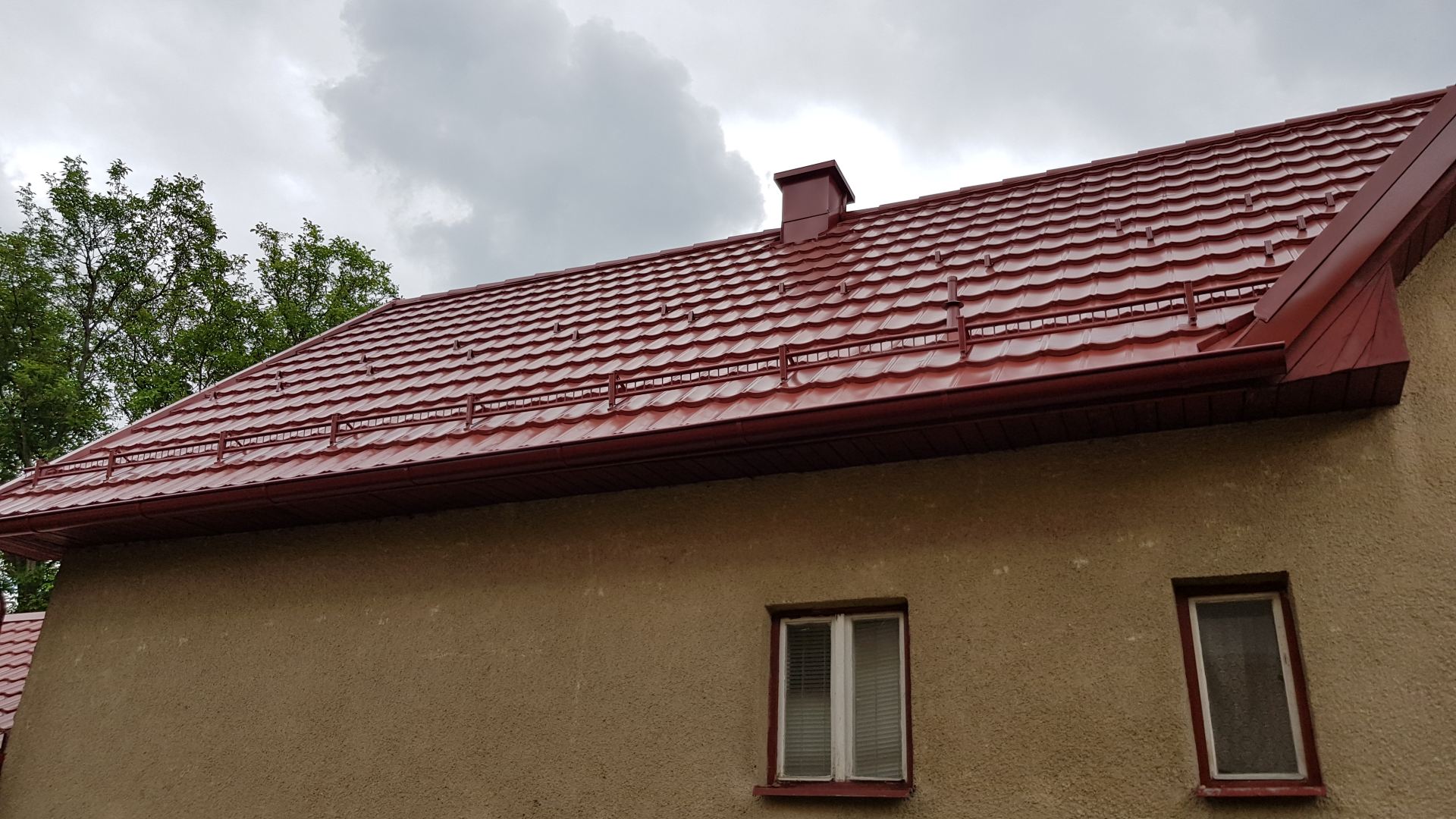 Červená plechová strecha s komínom a snehovou zábranou na béžovom dome, na pozadí zamračenej oblohy a stromov, vytvárajúca pokojný vzhľad.