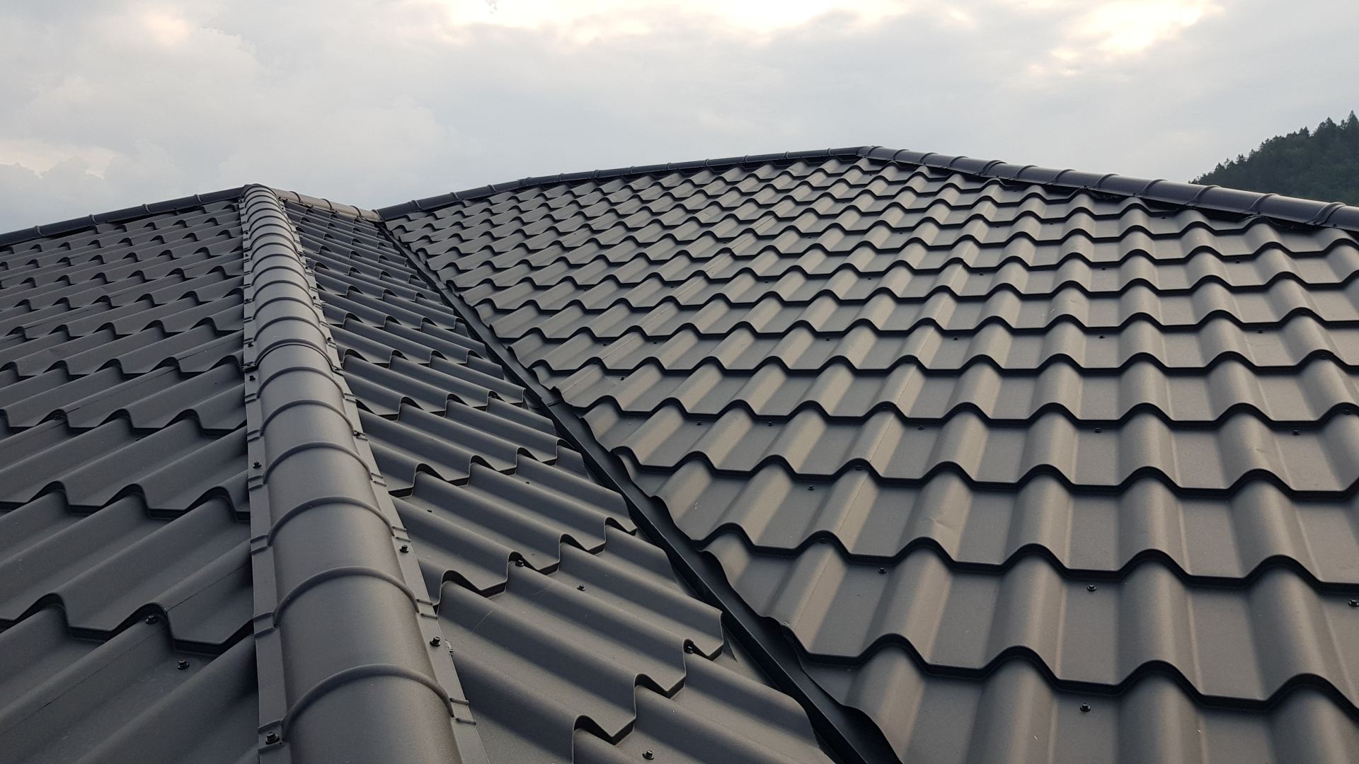 Detail tmavosivej plechovej strechy so škridlovým vzorom pod zamračenou oblohou. Vlnitý vzor vytvára jednotný a elegantný vzhľad a vyjadruje odolnosť.