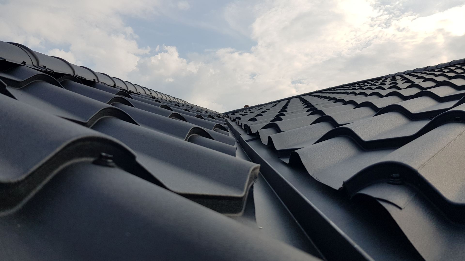 Detail modernej vlnitej plechovej strechy na pozadí čiastočne zamračenej oblohy. Elegantné tmavé strešné škridly vyvolávajú pocit odolnosti a štýlu.
