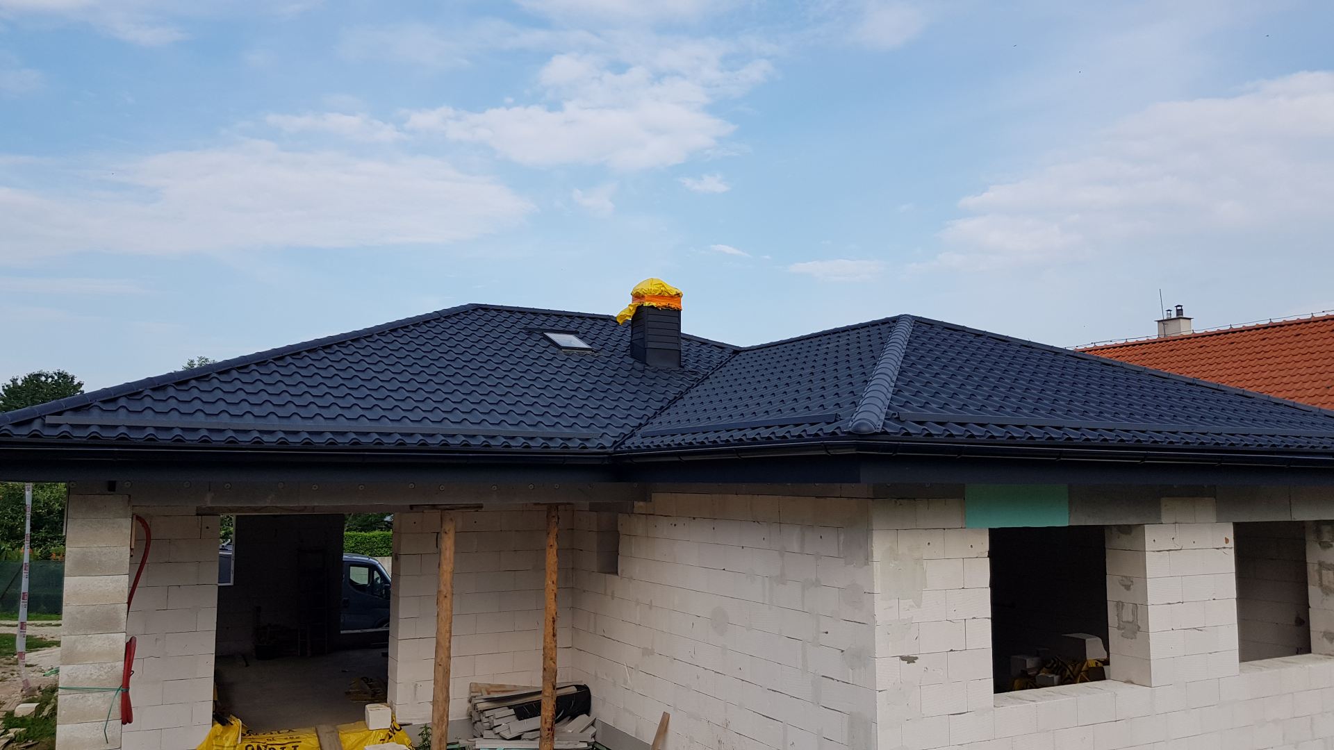 Novostavba so strechou z antracitovo sivej škridly na pozadí modrej oblohy. Nedokončený komín je prikrytý žltou plachtou.