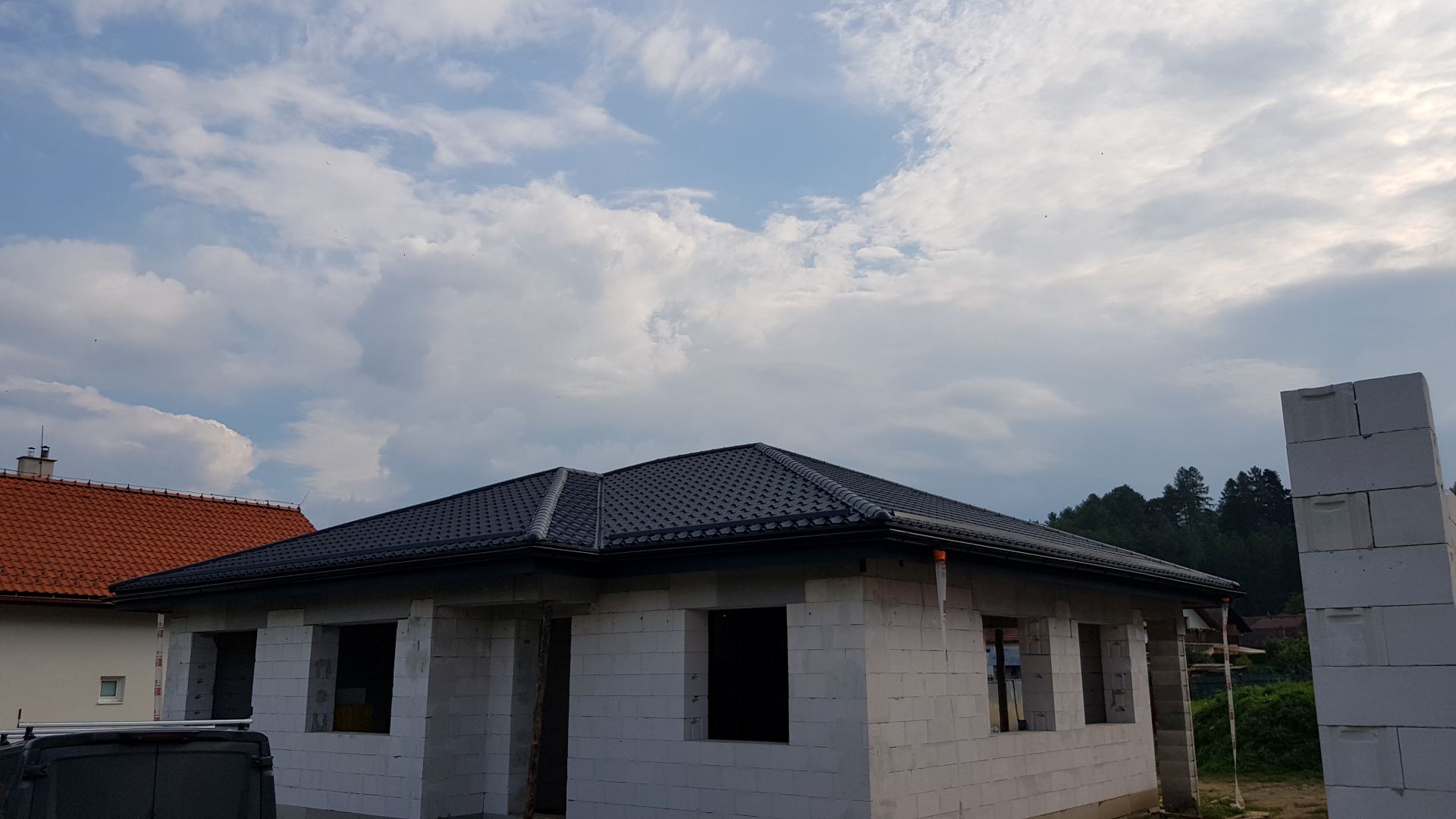 Prízemný dom vo výstavbe so sivými tehlovými múrmi a tmavou škridlovou strechou. Zamračená obloha s rozptýlenými oblakmi dodáva pokojnú, prechodnú atmosféru.