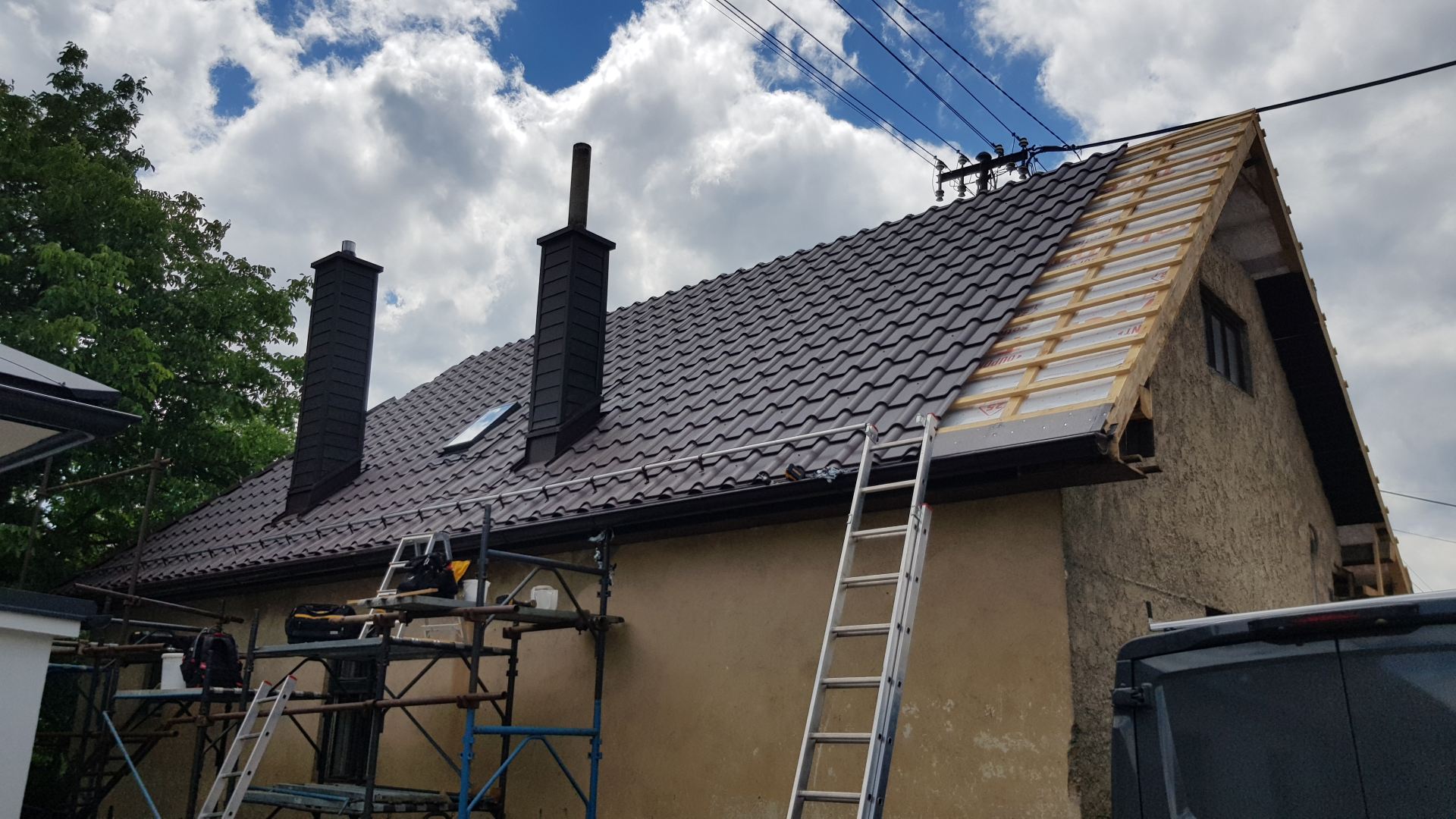 Strešné práce na dome s čiastočne položenou kovovou strechou, lešením a rebríkmi. V pozadí zamračená obloha a zelené stromy.
