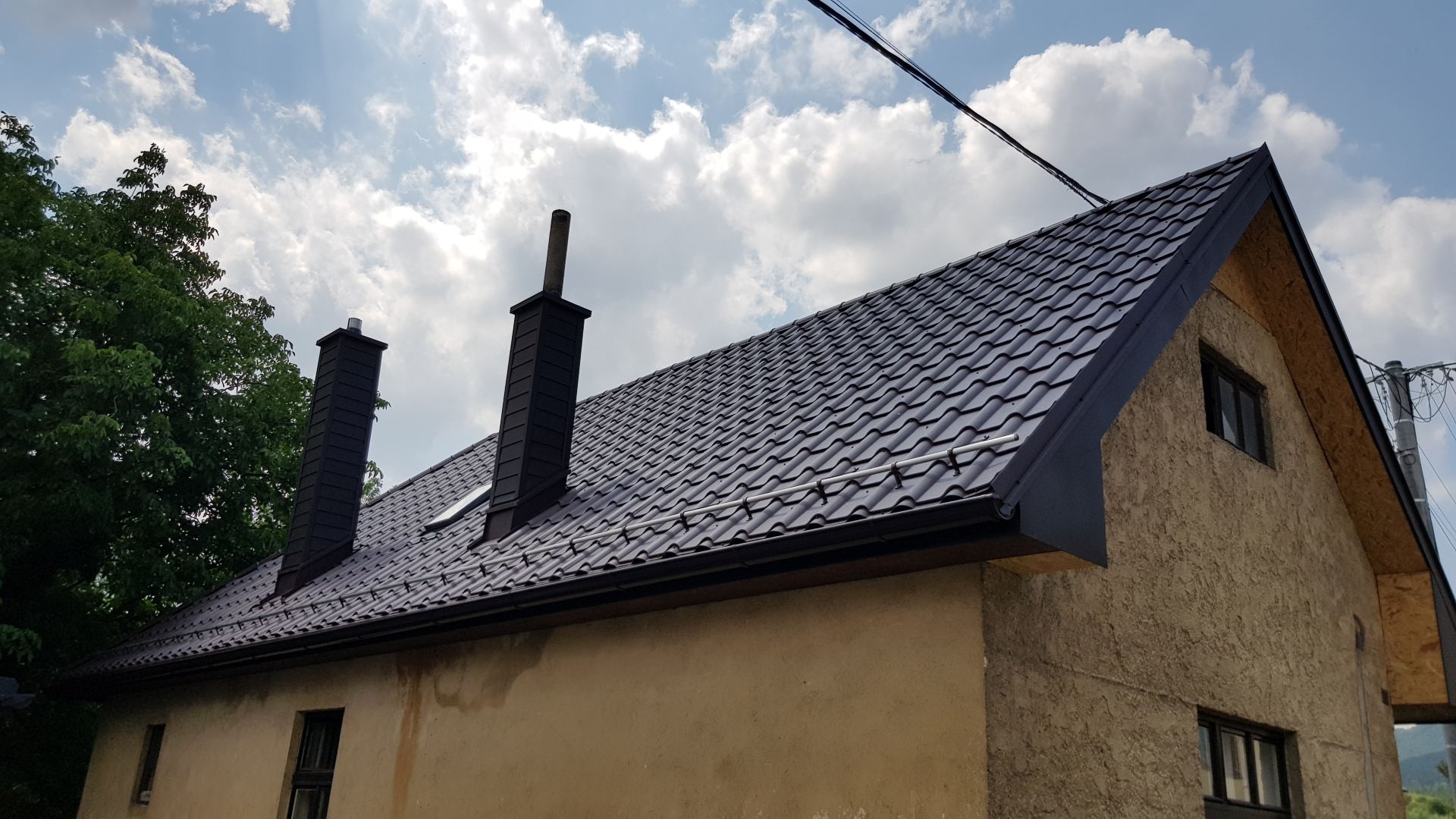 Rustikálny dom so štruktúrovanou béžovou stenou a tmavou plechovou strechou s dvoma komínmi. Obloha je čiastočne zamračená a naľavo je vidieť strom.