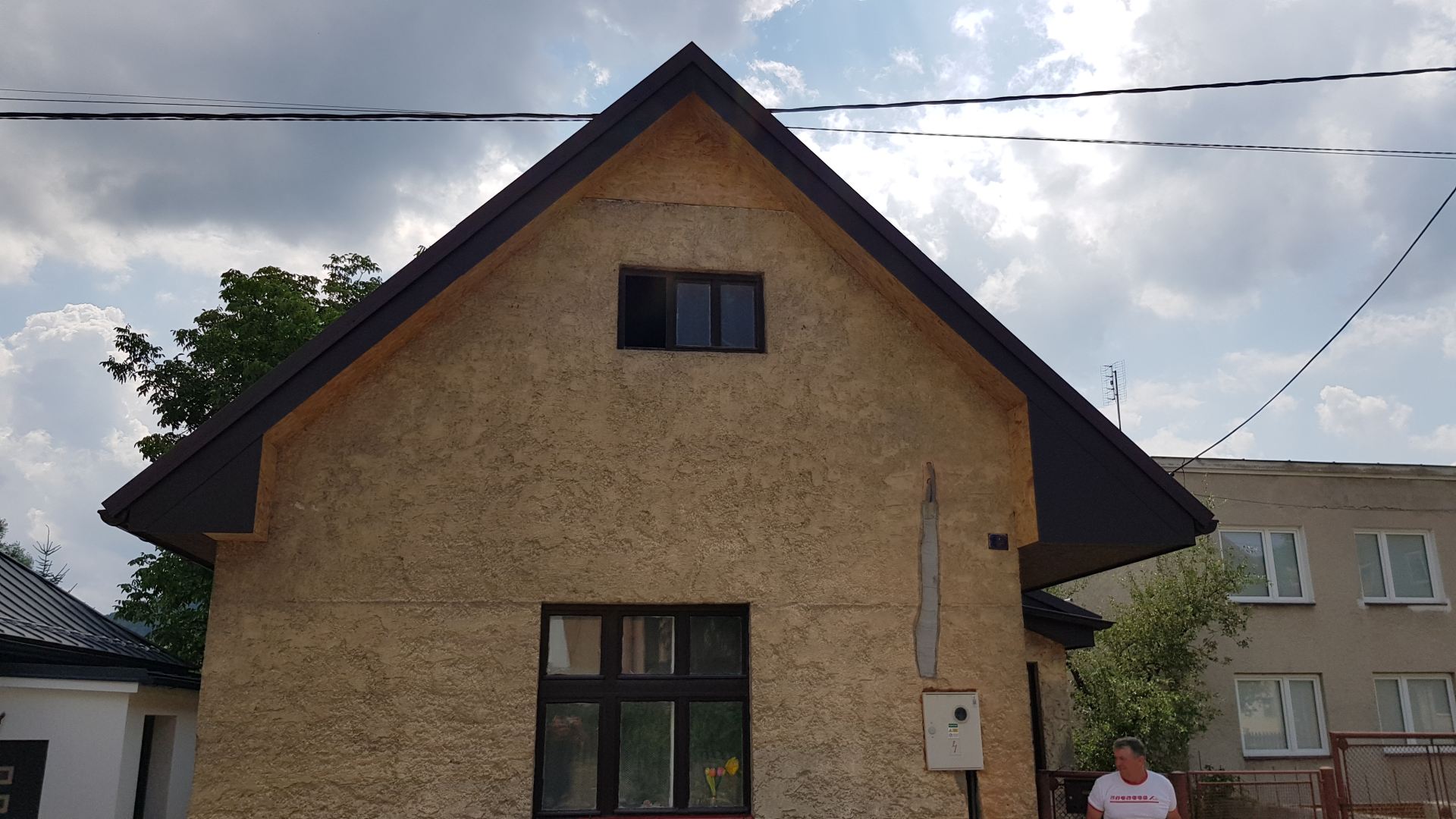 Béžový omietnutý dom s strmou tmavou strechou pod zamračenou oblohou. Sú tam dve okná a osoba stojí v pravom dolnom rohu.