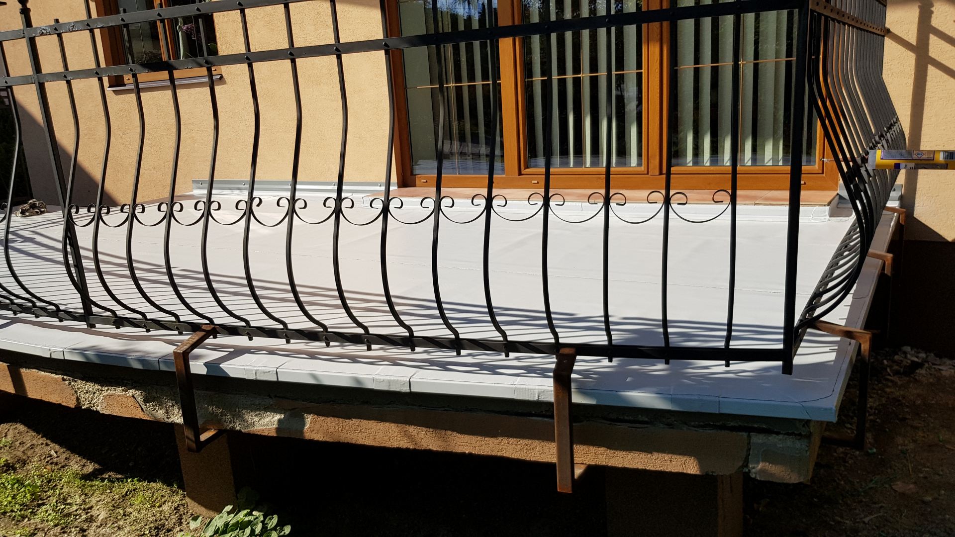 Zakřivené kovové zábradlie obklopuje malý balkón s béžovou stenou a oknami s drevenými rámami v pozadí. Slnečné svetlo vrhá tiene na bielu podlahu balkóna.