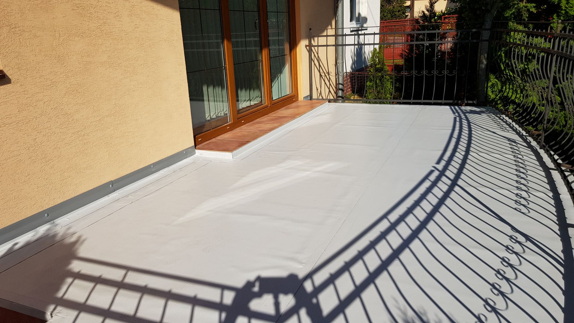 Slnečný balkón s bielou podlahou, béžovými stenami a sklenenými dverami. Čierne kovové zábradlie vrhá zložité zakrivené tiene na podlahu, čím pridáva eleganciu.