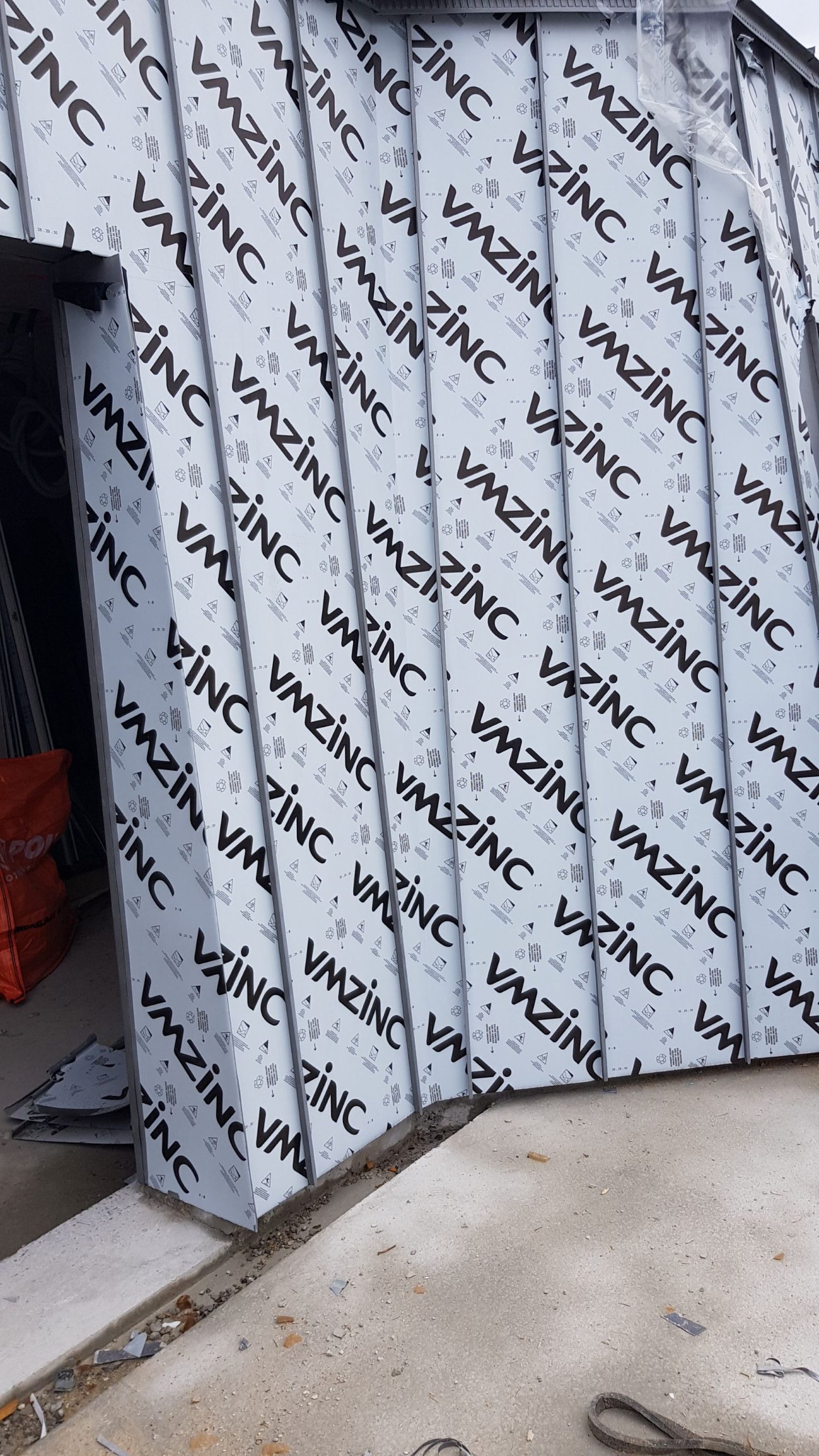 Zakrivené kovové panely s logom „VMZINC“ pokrývajú vonkajšiu časť budovy a prezentujú moderný architektonický dizajn. Vidno betónovú plochu a oranžový vak.
