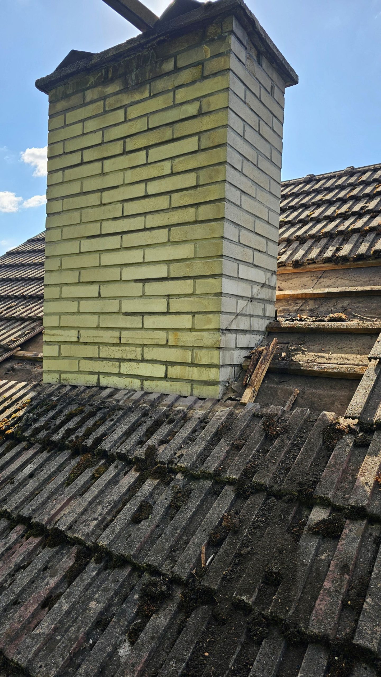 Starý tehlový komín na streche so zvetranými, machom pokrytými škridlami. Slnečné svetlo vrhá tiene, ktoré zvýrazňujú opotrebovaný stav komína. Nad ním modrá obloha.