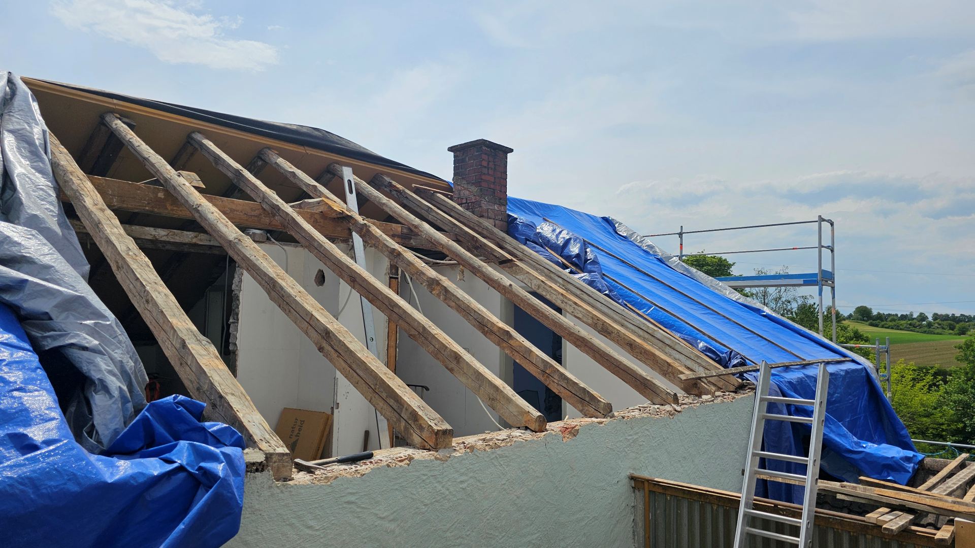 Čiastočne postavená strecha s odhalenými drevenými trámami a modrými plachtami, tehlový komín a rebrík na boku. V pozadí je vidieť vidiecku krajinu.