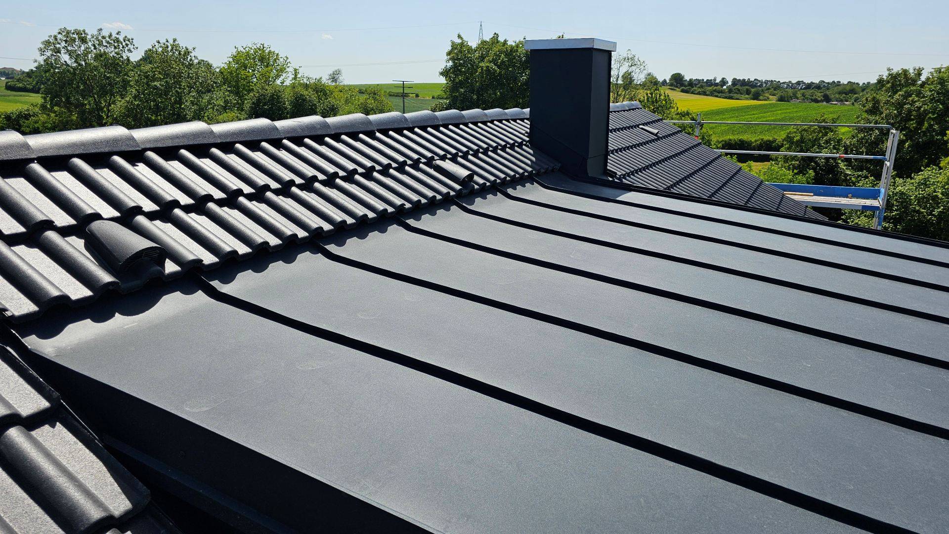 Elegantná moderná strecha s tmavými, hladkými škridlami a komínom v popredí. Okolie tvoria bujné zelené stromy pod jasnou modrou oblohou.